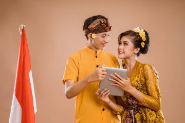 man and woman wearing balinese traditional clothing using tablet pc and smiling