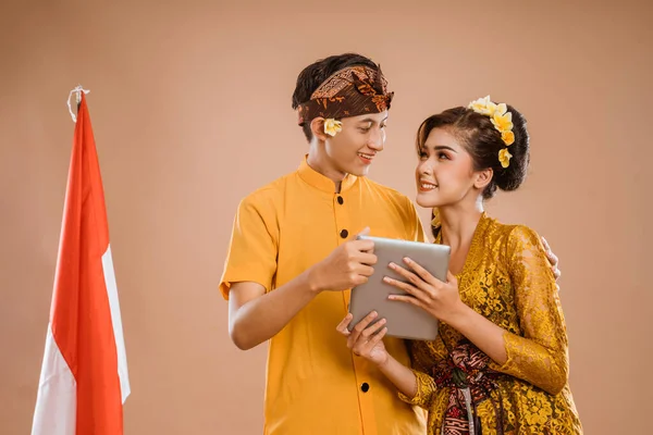 man and woman wearing balinese traditional clothing using tablet pc and smiling