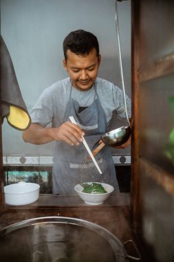 man seller stirring boiled noodles with chopsticks in a bowl on the cart