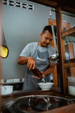 man seller in apron pouring vegetable oil from a bottle to bowl when preparing chicken noodles dish