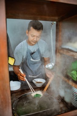 man seller draining boiled noodles with a drainer and chopsticks from the pan on the cart