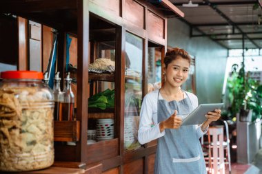 female chicken noodle vendor in apron smiling with thumbs up while using a pad in front of cart