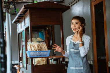 female salesperson in apron calling using smartphone with chicken noodle cart background