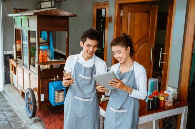 woman and man wearing apron using pad and cell phone together at chicken noodle stall