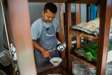 man seller in apron stir vegetable oil in the bowl when preparing chicken noodles dish