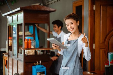 woman in apron smiling with thumbs up while holding pad at chicken noodle stall