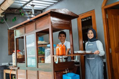 smiling two sellers in apron preparing chicken noodles dish with bowls on tray for customer