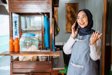 veiled woman wearing an apron talking on a cellphone while selling at a chicken noodle cart