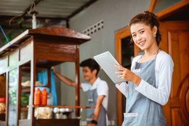 Asian woman in apron smiling while holding pad at chicken noodle stall