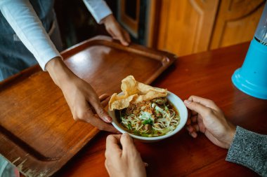 waiters hand serving a bowl of chicken noodle dish to customers on the wood table