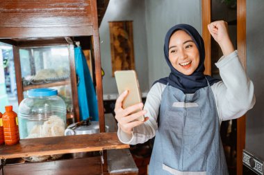 veiled woman wearing an apron happily looking at her cellphone while selling at a chicken noodle cart
