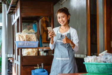 female chicken noodle seller in apron smiling with thumbs up while using mobile phone with cart background