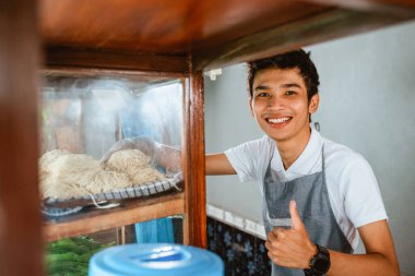 selling man wearing apron smiling with thumbs up while taking noodles from chicken noodle cart