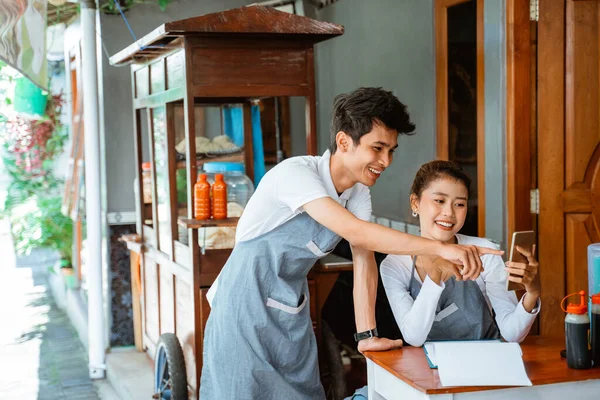 male and female sellers in apron using cell phones together at chicken noodle stall