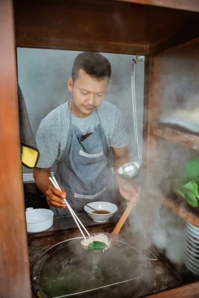 man seller draining boiled noodles with a drainer and chopsticks from the pan on the cart