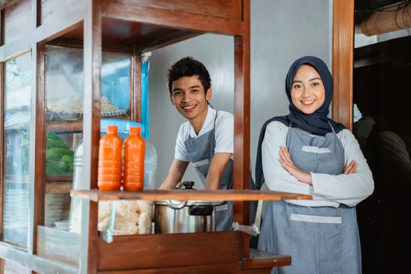 veiled woman wearing apron smiling with hands crossed and young man selling in chicken noodle cart