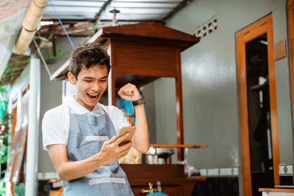 happy male merchant wearing apron looking at mobile phone screen with cart background