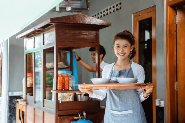 smiling female waitress carrying a bowl with a tray with a chicken noodle cart seller in the background