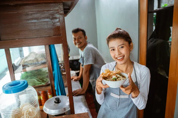 female seller smiling while bringing a bowl of chicken noodles for customers in a stall cart