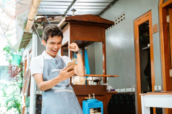 excited male salesman wearing apron when looking at mobile phone with cart background