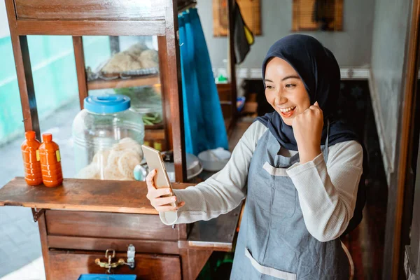 excited woman wearing apron looking at cell phone while selling in chicken noodle cart