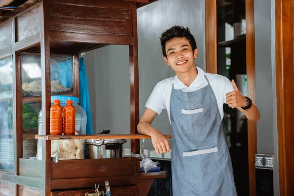 smiling young man wearing apron with thumbs up standing beside chicken noodle cart