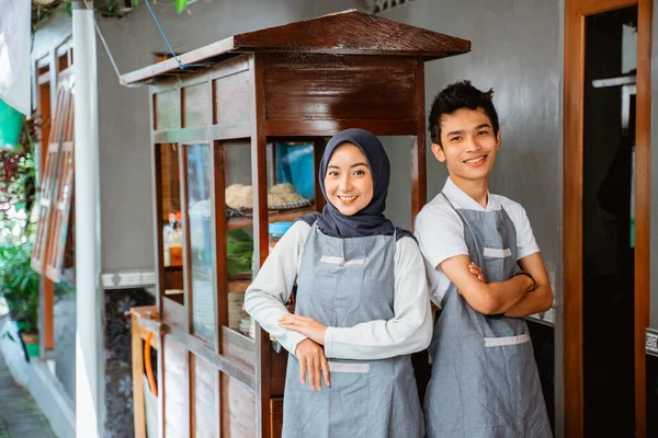 a couple of sellers wearing an apron with their hands crossed smiling while selling on a chicken noodle cart