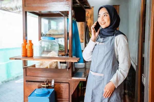 woman wearing an apron in a veil makes a cell phone call while selling at a chicken noodle cart