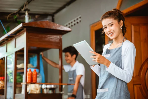 Asian woman in apron smiling while holding pad at chicken noodle stall