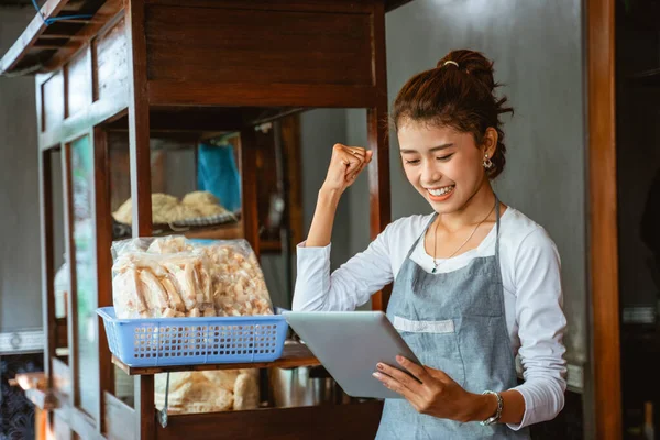excited saleswoman in apron with clenched hands while using tablet with chicken noodle cart background