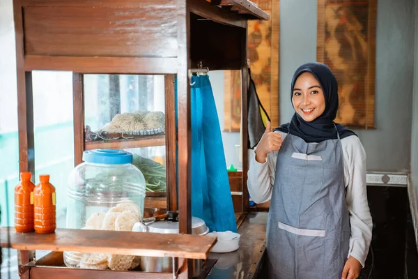 hijab woman wearing apron smiling while standing with thumbs up behind cart