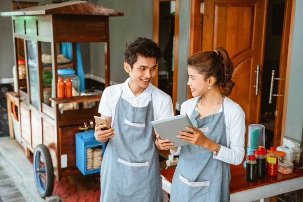 man and woman in apron holding pad and cell phone at chicken noodle stall