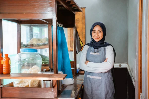 hijab woman wearing apron smiling while standing with crossed hands behind cart