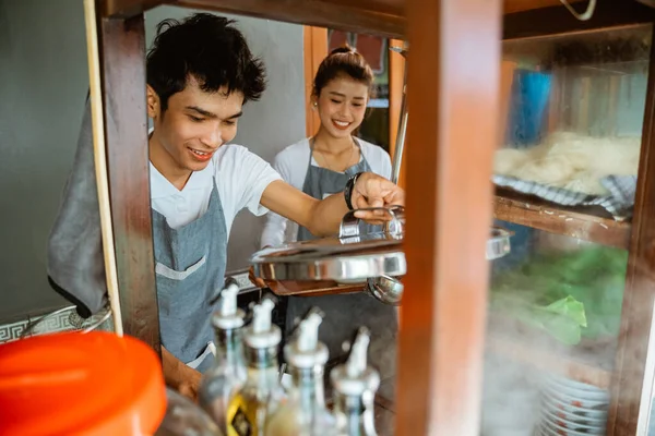 a man selling noodles cooks and holds a pot lid with the help of a woman when selling in a stall