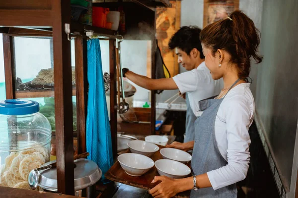 woman carrying an empty bowl with a tray while helping a man cook chicken noodles at a stall