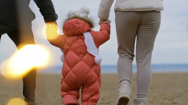 Anne ve baba bebeğe yürümeyi öğretiyor. Aile sahilde yürüyor.