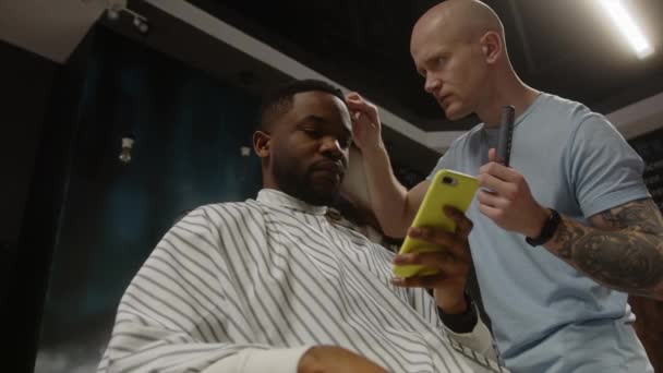 Лысый парикмахер расчесывает волосы афроамериканского клиента, который пользуется смартфоном — стоковое видео