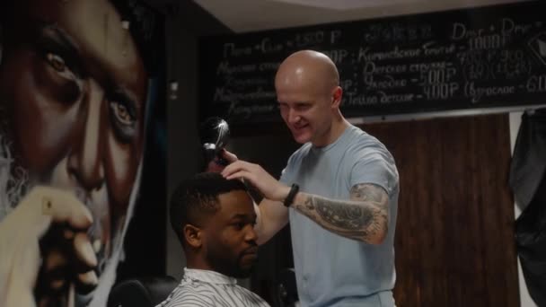 Un peluquero calvo seca la cabeza de un cliente de peluquería negro con un secador de pelo — Vídeos de Stock