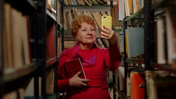 Een oudere vrouw maakt een foto met boeken in de bibliotheek. Selfie voor sociale netwerken — Stockfoto