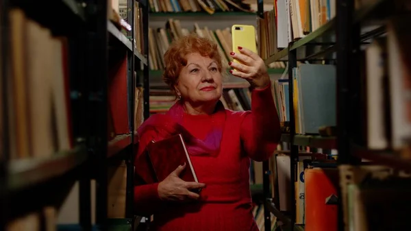 Yaşlı bir kadın kütüphanede kitaplarla fotoğraf çekiyor. Sosyal ağlar için selfie