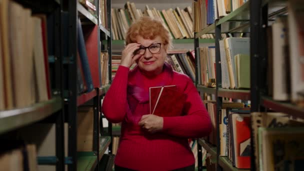 Μια ηλικιωμένη γυναίκα με γυαλιά στέκεται στη βιβλιοθήκη ανάμεσα στα ράφια με τα βιβλία στα χέρια της. — Αρχείο Βίντεο