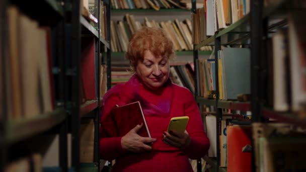 Seorang wanita tua mengambil foto dengan buku di perpustakaan. Selfie untuk jejaring sosial — Stok Video