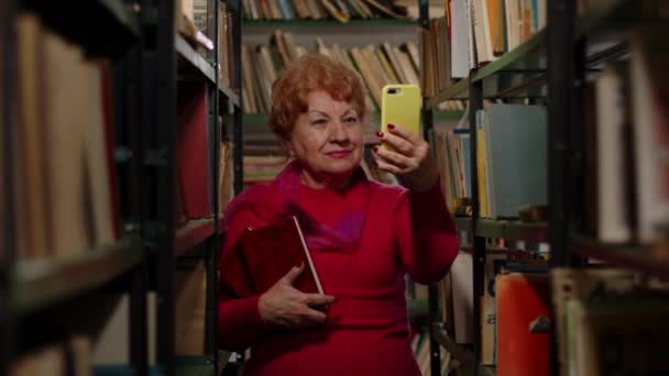 Una donna anziana scatta una foto con dei libri in biblioteca. Selfie per i social network — Video Stock