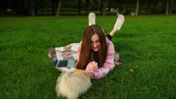 En flicka leker och skrattar med en rolig liten hund liggande på gräset på en filt i parken — Stockvideo