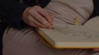 Ressam, oturma odasındaki kanepede oturan sarı kağıt bir eskiz defterine bir kız resmi çiziyor.