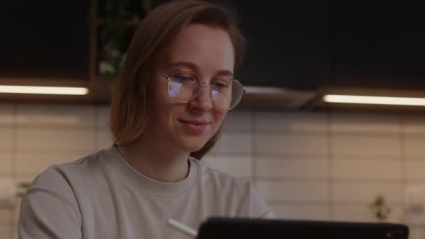 Усміхнена дівчина в окулярах навчається вдома на кухні за допомогою планшета і цифрової ручки — стокове відео