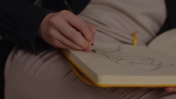 De kunstenaar tekent een schets van een meisje in een geel papieren schetsboek zittend op een bank in de woonkamer — Stockvideo