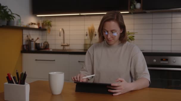 Una talentosa artista del concepto de chica dibuja en una tableta con una pantalla digital un proyecto de un futuro juego casual en 3D, sentado en una mesa en casa en la sala de estar — Vídeos de Stock