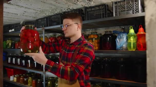 En jordbrukare med glasögon kontrollerar kvaliteten på pickles i källaren. Konserverad mat i källaren — Stockvideo