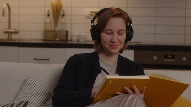 Όμορφο κορίτσι κάθεται στον καναπέ, ακούγοντας μουσική στα ακουστικά και ζωγραφίζοντας σκίτσα σε ένα ψηφιακό tablet — Αρχείο Βίντεο